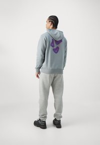 Lys grå hettegenser med et lilla abstrakt design på ryggen, kombinert med grå joggebukser og svarte joggesko. Uformell og komfortabel passform.