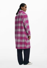 Lungo cappotto a quadri in rosa e grigio, realizzato in lana, con colletto e spacchi laterali. Abbinato a pantaloni larghi blu navy e scarpe bianche.
