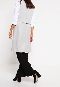 Grå ullväst med skräddarsydd design, bälte på baksidan, lagd över en vit skjorta och svarta vida byxor, tillsammans med svarta klackskor.