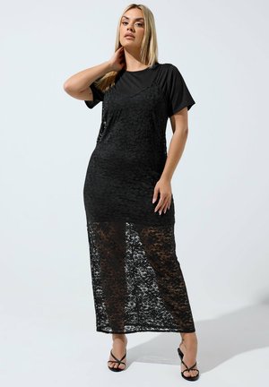 Vrouw poseert in een zwart kanten mouwloze jurk, gelaagd over een zwart shirt met korte mouwen, met zwarte sandaalhakken tegen een effen achtergrond.