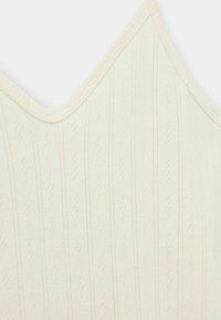 Krém színű pántos felső V nyakkivágással. Jellemzője a függőleges csíkok és kis gyémántformák textúrájú mintázata. Puha anyag.