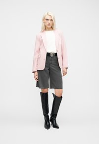 Νεαρή γυναίκα που φοράει ροζ σακάκι, λευκό πουκάμισο, μαύρες μπότες μέχρι το γόνατο και μαύρα τζιν σορτς με ζώνη που έχει αγκράφα σε σχήμα καρδιάς, στέκεται μπροστά σε λευκό φόντο.