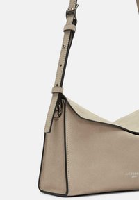 Beige Wildleder-Handtasche mit schwarzem Besatz, ausgestattet mit einem langen verstellbaren Schultergurt und einem schrägen Oberklappendesign. Minimalistische Markenkennzeichnung auf der Vorderseite.