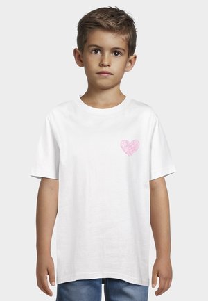 Chłopiec o krótkich włosach, ubrany w białą koszulkę z małym różowym sercem na lewej piersi, stojący na tle jednolitego szarego tła.