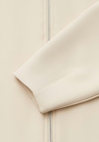 Close-up van een beige jas met een gladde textuur, een rits aan de linkerkant en een omgeslagen manchet met nette stikdetails.