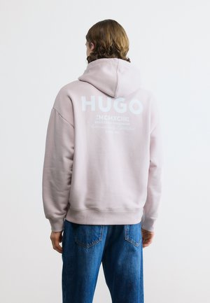 Person trägt einen hellrosa Hoodie mit dem Schriftzug "HUGO" und römischen Zahlen auf dem Rücken, kombiniert mit blauen Denim-Jeans, von hinten gesehen.