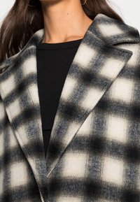 Kostkovaný vlněný kabát v černé a bílé barvě, se širokým límcem, měkkou texturou a uvolněným střihem. Vzor se skládá z velkých kontrastních čtverců.