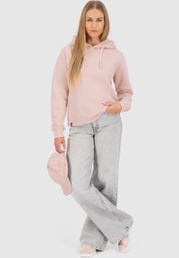 Felpa a cappuccio rosa chiaro con cordini, abbinata a jeans larghi grigi chiari. Un cappellino rosa coordinato tenuto in una mano.