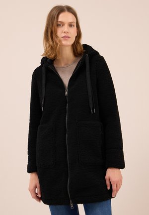 Femme portant une veste à capuche zippée noire en peluche avec de grandes poches avant sur une chemise beige et un jean bleu, sur un fond neutre.