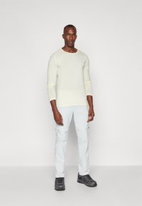 Haut à manches longues crème clair associé à un pantalon cargo gris clair. L'ensemble est complété par des chaussures gris foncé. Texture lisse, design minimal.