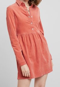 Šedohnědé kordurové šaty košilového typu s dlouhými rukávy, knoflíkovým zapínáním vpředu a nařaseným pasem. Hladká textura a jemný lesk.