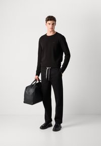 Sort crew-neck sweater, sorte bukser med snørre, bærer en sort lædertaske. Tekstureret stof med minimal branding.