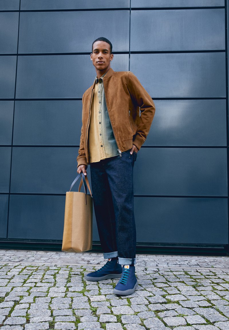 Brun suede bomberjakke, lys blå skjorte med knapper, mørke denimbukser, blå sneakers og en brun papirpose mod en grå væg.