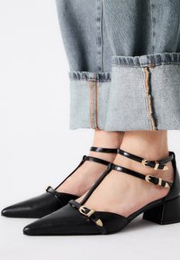 Zapatos de cuero negro con punta afilada y diseño de correa en T. Presentan tres correas ajustables con hebillas doradas y un tacón bajo y cuadrado.