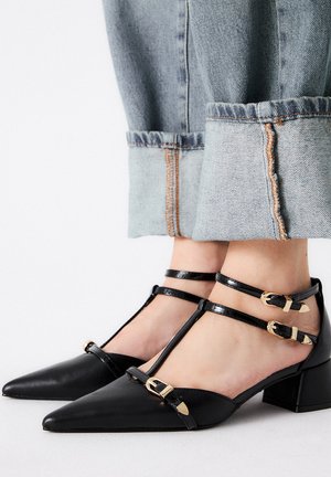 Zapatos de cuero negro con punta afilada y diseño de correa en T. Presentan tres correas ajustables con hebillas doradas y un tacón bajo y cuadrado.