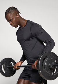 Fekete hosszú ujjú sportfelső, texturált anyagból, kerek nyakkivágással, amelyet egy fekete súlytárcsa felett tartanak egy emelés közben.