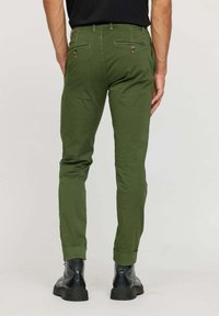 Pantalons en coton verts avec une coupe slim, dotés de poches arrière avec fermetures à bouton. Ourlets retroussés et portés avec des bottes en cuir noires.