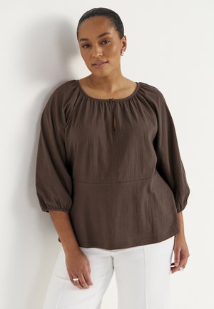 Kobieta o ciemnych włosach, w brązowej bluzce z długim rękawem i marszczonym dekoltem, stoi przy białej ścianie, ubrana w białe spodnie.