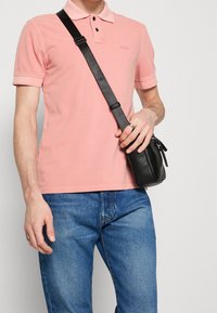 Chemise polo rose avec un col à deux boutons, associée à un jean en denim bleu. Un petit sac à bandoulière noir est porté sur l'épaule.