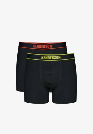 Hemdloser Mann mit kurzen Haaren, der schwarze Henderson-Boxershorts trägt, steht mit vor dem Körper verschränkten Händen vor einem schlichten hellen Hintergrund.