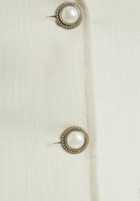 Witte stof met een gestructureerd patroon, met een zilverkleurige knop met een parel in het midden en omliggende versieringen.