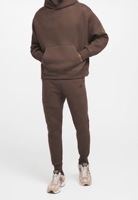 Sweatshirt marron avec une capuche et une poche, associé à un jogging assorti avec un motif texturé. Porté avec des baskets beiges.