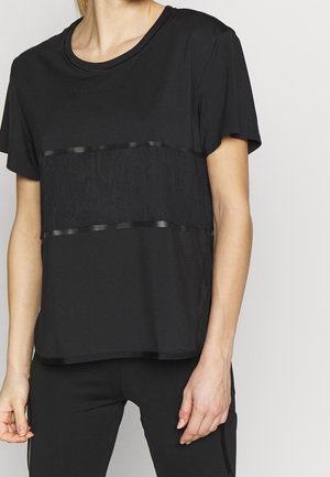 Mujer con camiseta negra de manga corta con un panel texturizado horizontal en el pecho y pantalones negros lisos.