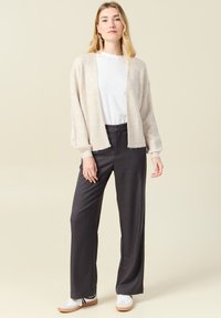 Ljusbeige cardigan i mjukt, strukturerat material med öppen front, kombinerad med mörkgrå vida byxor och vita sneakers.