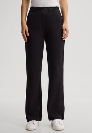 Woman wearing black wide-leg pants with a textured waistband and white sneakers, standing against a plain background.