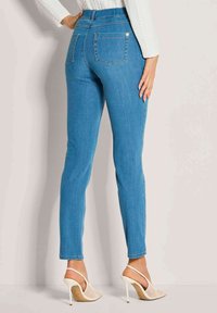 Frau in hellblauen, eng anliegenden Jeans und weißen, hochhackigen Netz-Slingback-Schuhen, steht mit einer Hand auf der Hüfte.