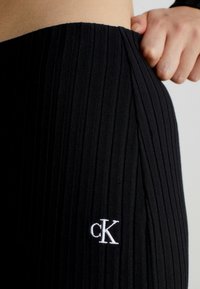 Pantalones negros de canalé con textura vertical, que presentan un logo bordado en blanco "cK" en el lateral. La tela parece elástica y suave.