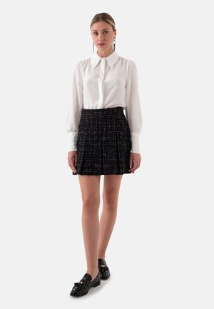 Witte, met kraag button-up blouse gecombineerd met een geplooide zwarte rok met een gestructureerd ruitpatroon, vergezeld van glanzende zwarte loafers.