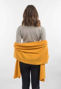 Gul fleece-sjal draperet over skuldrene, blød tekstur, lagdelt design, kontrast til grå top, parret med sorte bukser.