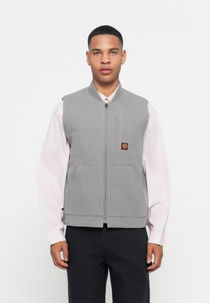 Man wearing light pink long-sleeve shirt under a gray zip-up vest with a small colorful patch, paired with black pants, against a white background.