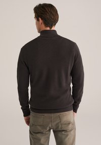 Dunkelgrauer Strickpullover mit hohem Kragen und gerippten Bündchen. Kombiniert mit hellbraunen, strukturierten Hosen mit Gesäßtaschen.