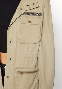 Khaki pracovní bunda s texturovaným okrajem kapes, se zapínáním na patentky a několika kapsami, včetně jedné na zip. Má jednoduchý design.