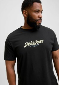 Fekete pamut póló, elöl krém és kék színű grafikával, amelyen a "JACK&JONES WORLDWIDE STUDIO" felirat szerepel, klasszikus kerek nyakkivágással.
