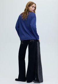 Vrouw met golvend haar, gekleed in een blauw gestreept shirt met lange mouwen en zwarte wijde broek met leren zakken met studs, die een grijze sjaal vasthoudt.