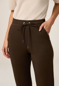 Zbliżenie na osobę w ciemnobrązowych spodniach z gumką w pasie i jedną ręką w kieszeni, w zestawieniu z jasnobeżową bluzką.