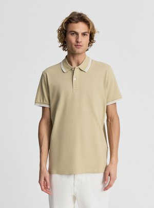 Młody mężczyzna z kręconymi blond włosami, ubrany w beżową koszulkę polo z białymi lamówkami i białe spodnie, stojący na jednolitym tle.