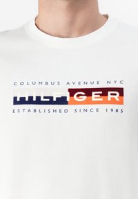 White cotton t-shirt featuring a multicolored graphic with the text “COLUMBUS AVENUE NYC HILFIGER ESTABLISHED SINCE 1985” in bold font.