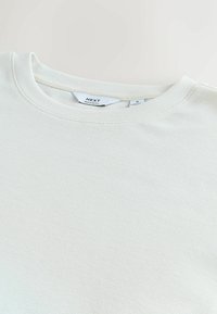 Tricou alb texturat cu un design rib onat, decolteu rotund și cusături vizibile. Eticheta este vizibilă, indicând "NEXT" și mărimea "M."