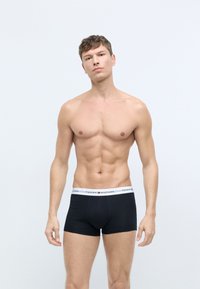 Sorte boxer briefs med en hvid elastik, der viser "TOMMY HILFIGER." Materialet ser glat og strækbart ud. Modellen står med armene afslappet.