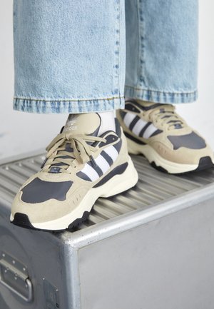 Osoba nosząca beżowo-czarne buty sportowe Adidas z białymi paskami i jasnoniebieskie dżinsy, stojąca na metalicznej powierzchni.