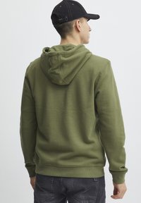 Sudadera verde oliva con capucha ajustable, de textura suave, con puños y dobladillo acanalados. Combinada con pantalones cargo gris oscuro.
