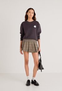 Hanorac gri închis cu un logo alb, combinat cu o fustă plisată în carouri în nuanțe de pământ. Modelul poartă pantofi loafer negri și șosete albe.