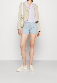 Žena v béžovém krátkém blejzru přes lila košili na knoflíky, světle modrých džínových kraťasech, krémových mokasínách a s krémovou kabelkou přes rameno.