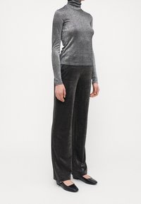 Glänzendes schwarzes Rollkragenoberteil und weite Hosen aus einem strukturierten Stoff; dunkles, metallisches Aussehen; kombiniert mit schwarzen flachen Schuhen.