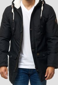 Veste matelassée noire avec une capuche doublée en fausse fourrure, fermeture éclair et boutons, plusieurs poches avant, et cordons de serrage contrastants.