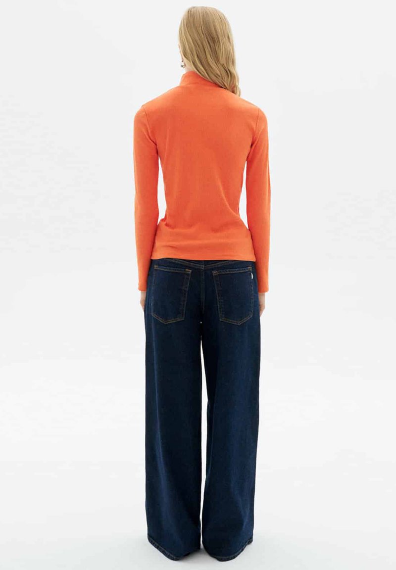 Oranges, geripptes Langarmoberteil, kombiniert mit weiten, dunkelblauen Jeans. Das Outfit wird von hinten gezeigt und zeigt sowohl schmale als auch lockere Elemente.